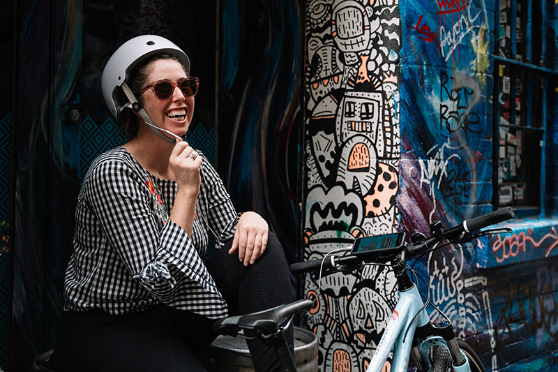 Woman sits next to a bike, holding microphone and laughing while participating in Rider Spoke.